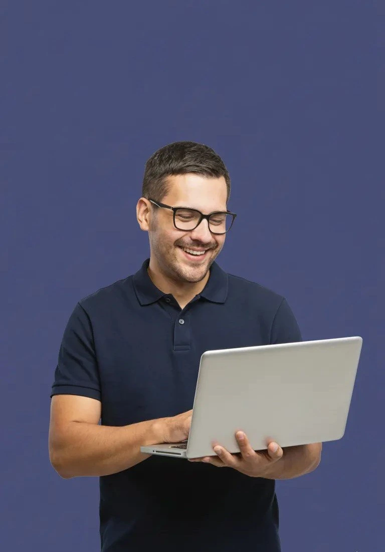 Smiling man wearing glasses using a laptop while standing against a blue background.